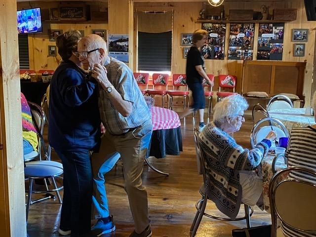 People dancing and seated in a warmly lit diner; a man and woman embrace on the dance floor, others watch and dine.