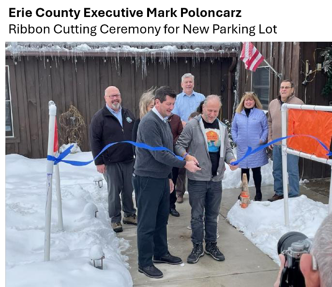 Erie County Executive Mark Poloncarz cuts blue ribbon for new parking lot, snowy setting.