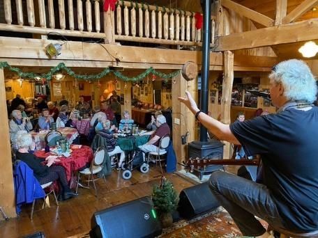 Musician on stage performing for seated audience in a rustic lodge decorated for a holiday.