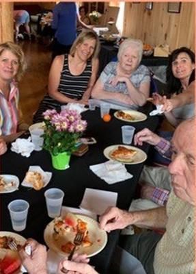 Group of people eating at a table with pizza, drinks, and a flower pot indoors.