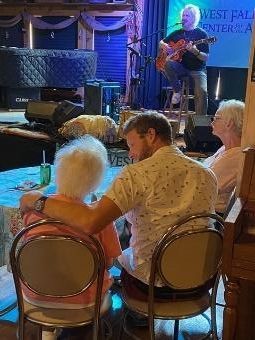 Man comforts older woman at a live music venue; guitarist performs onstage.