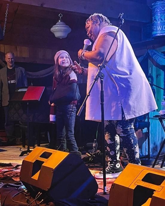 A performer with a microphone speaks to a child on stage; a band member stands behind a keyboard.