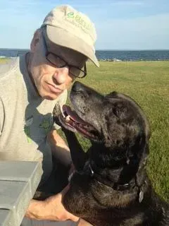 Man in glasses and cap pets a black dog outdoors, near water.