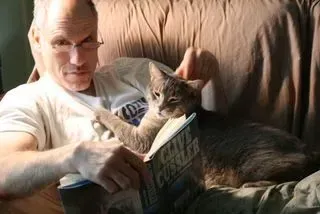 Man in glasses reading with cat resting on book in a chair.