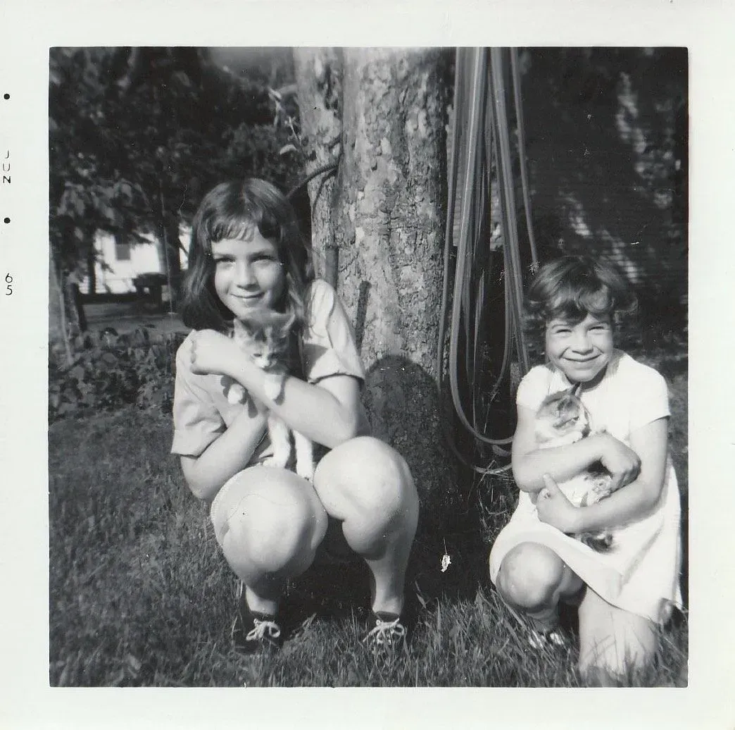 Two young girls squatting on grass, each holding a kitten.