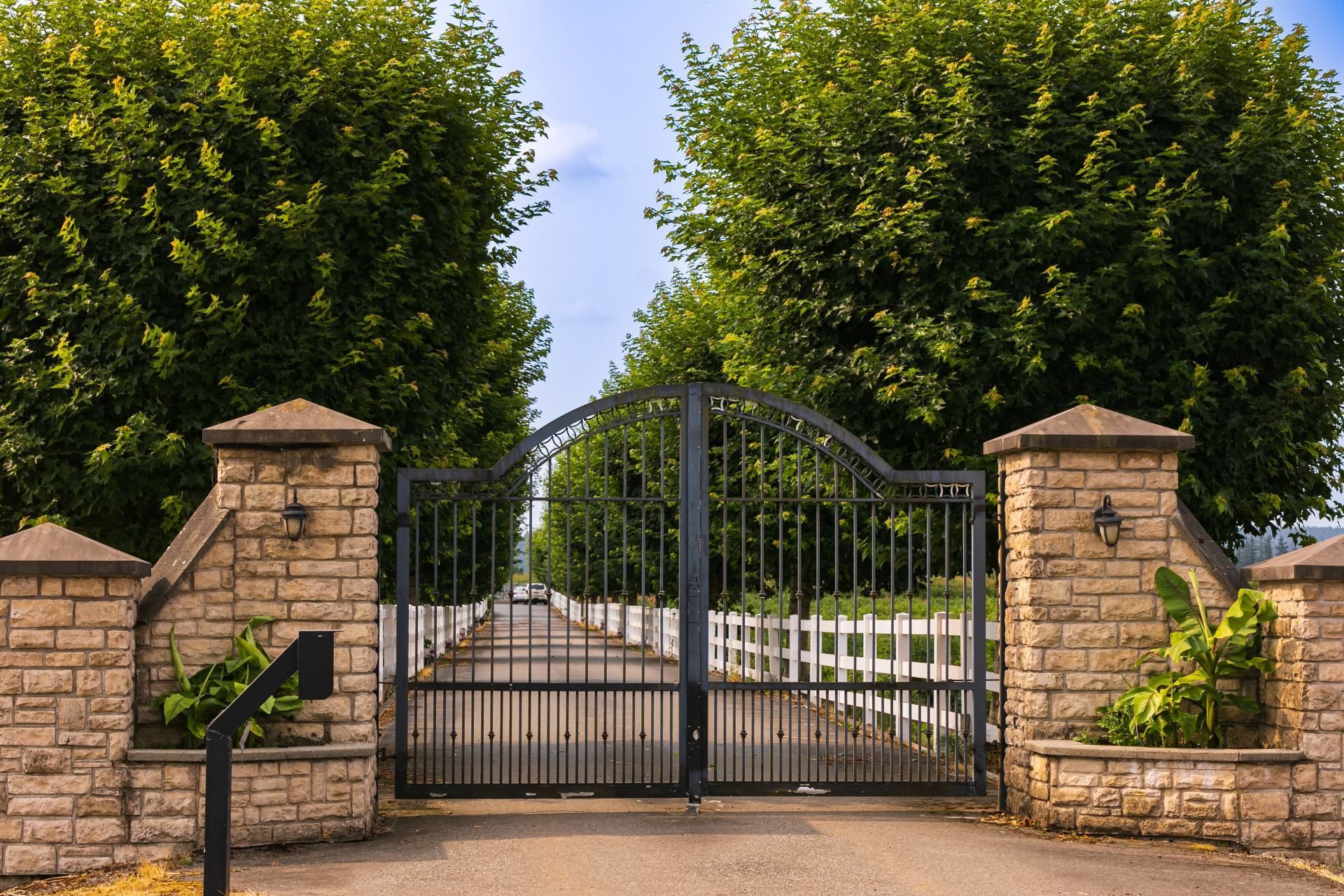 Stone pillars frame a black wrought iron gate on a paved road, leading to a white fence and trees.