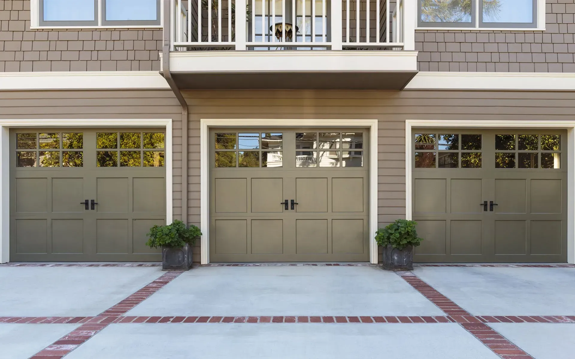 Three olive-green garage doors with windows, brick accents, and potted plants in front of a two-story building.