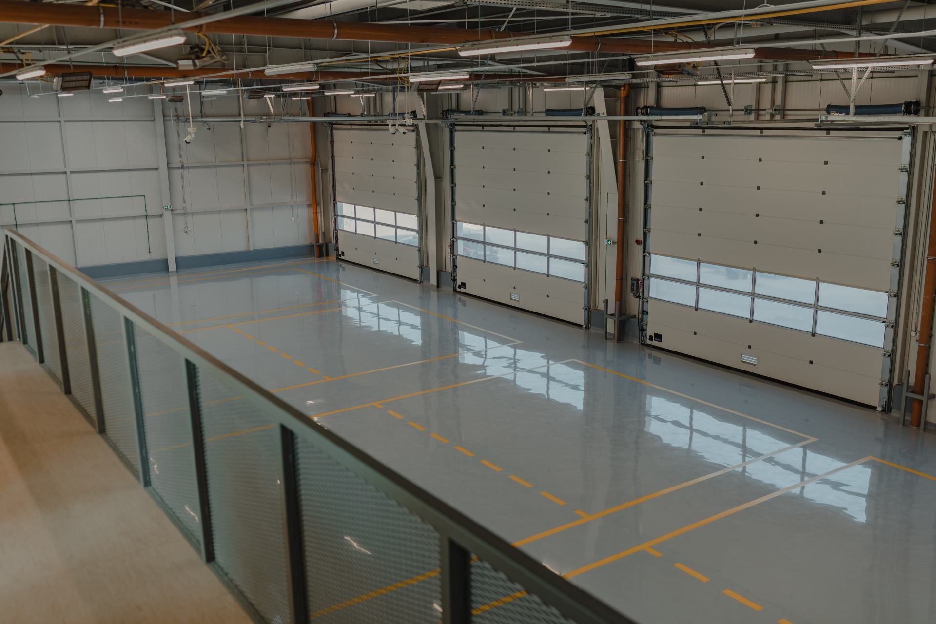 Warehouse interior with three closed loading dock doors. Grey floor, metal railing, overhead lights.