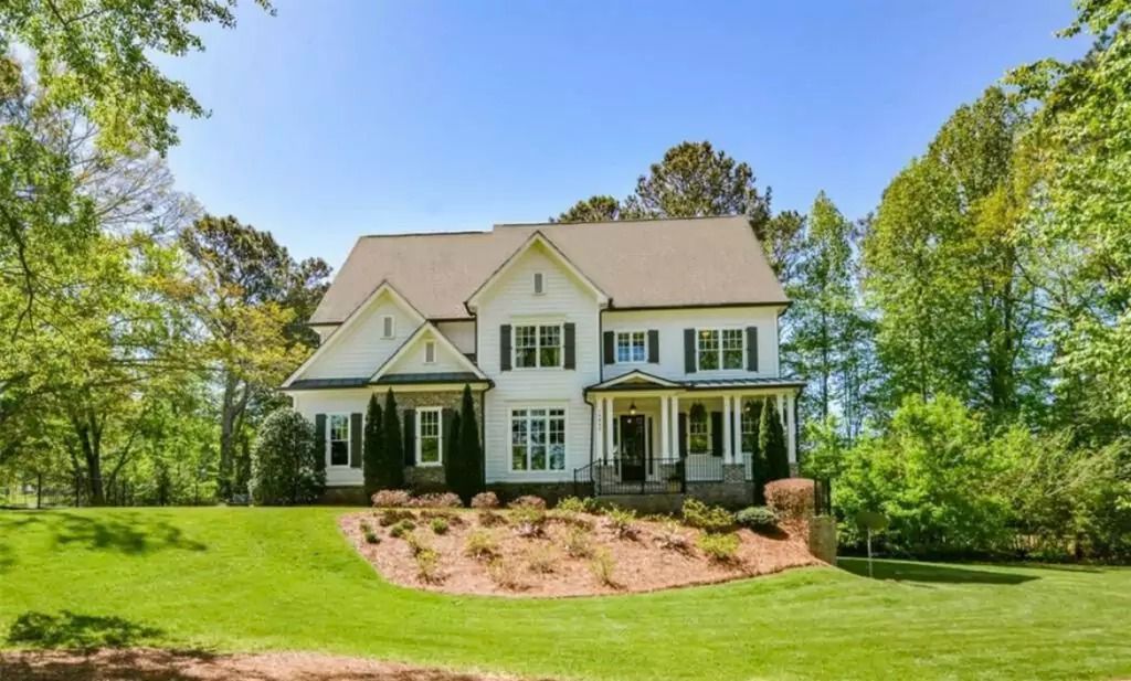 White two-story house with black shutters, on a green lawn with landscaping; trees in the background under a blue sky.