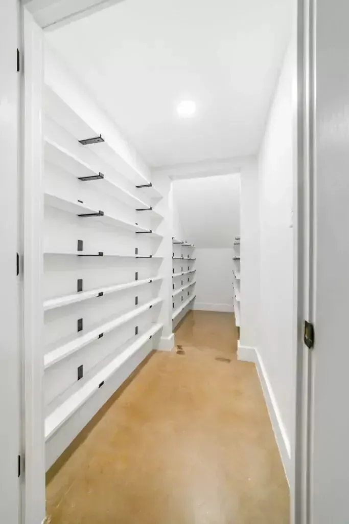 White pantry with long shelves, brown floor, and a bright overhead light.