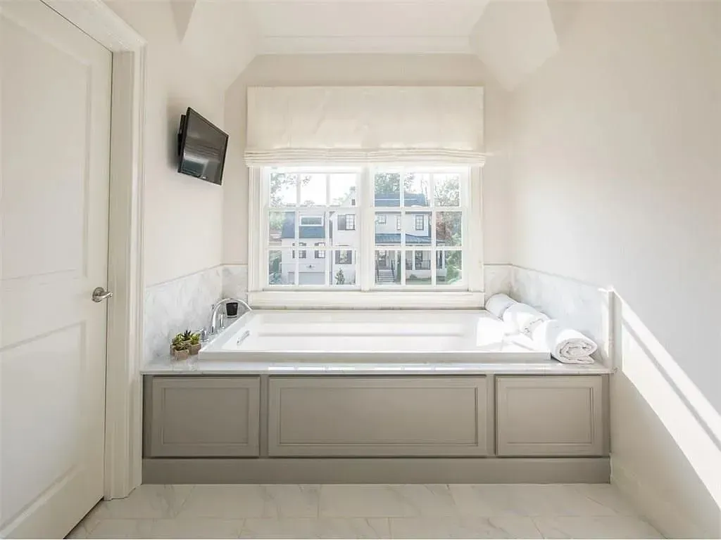 Bathroom with tub under a window. Gray cabinetry, white walls and marble, a TV, and a white door.