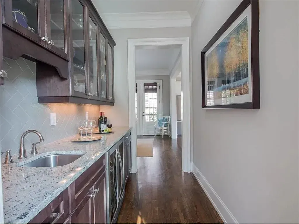 A home wet bar with dark wood cabinets, light granite countertops, a small sink, and glass-front cabinets in a hallway.
