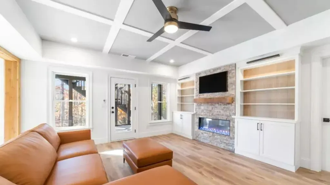 A modern living room with a tan leather sofa, stone fireplace, white built-in bookshelves, and a coffered ceiling.