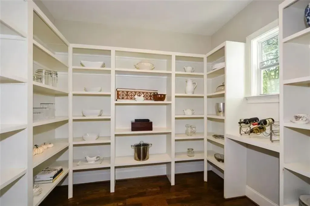 White pantry with many shelves, holding dishes and kitchen items. Dark wooden floor.