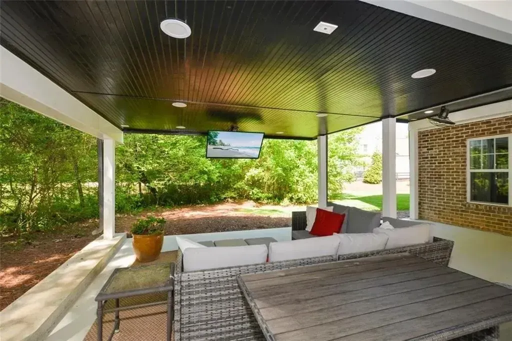 Covered patio with sectional sofa, TV, and brick floor. Dark ceiling, view of green trees.