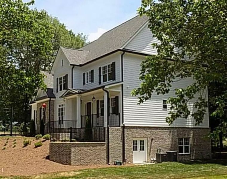 Two-story white house with brick foundation, black shutters, and porch under a blue sky, surrounded by trees.