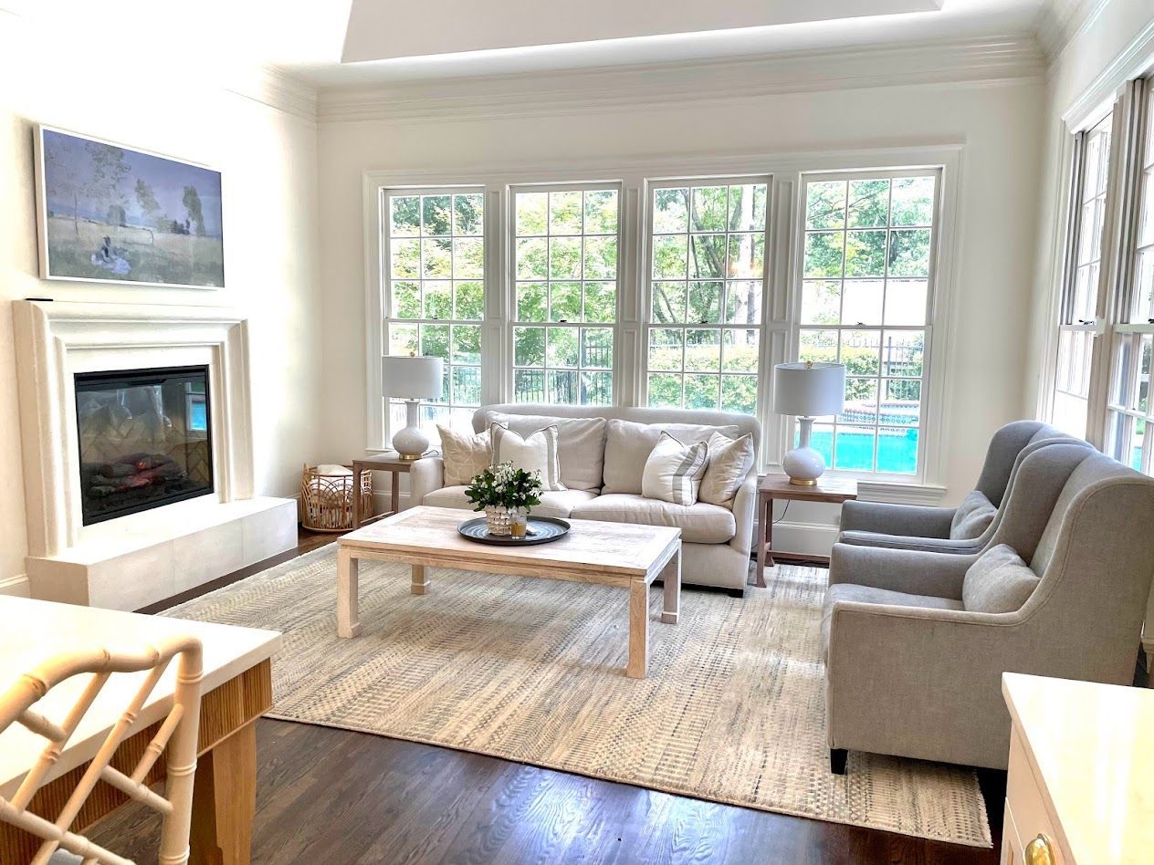Living room with a fireplace, windows, and light-colored furniture, including a sofa and armchairs, with a large rug.