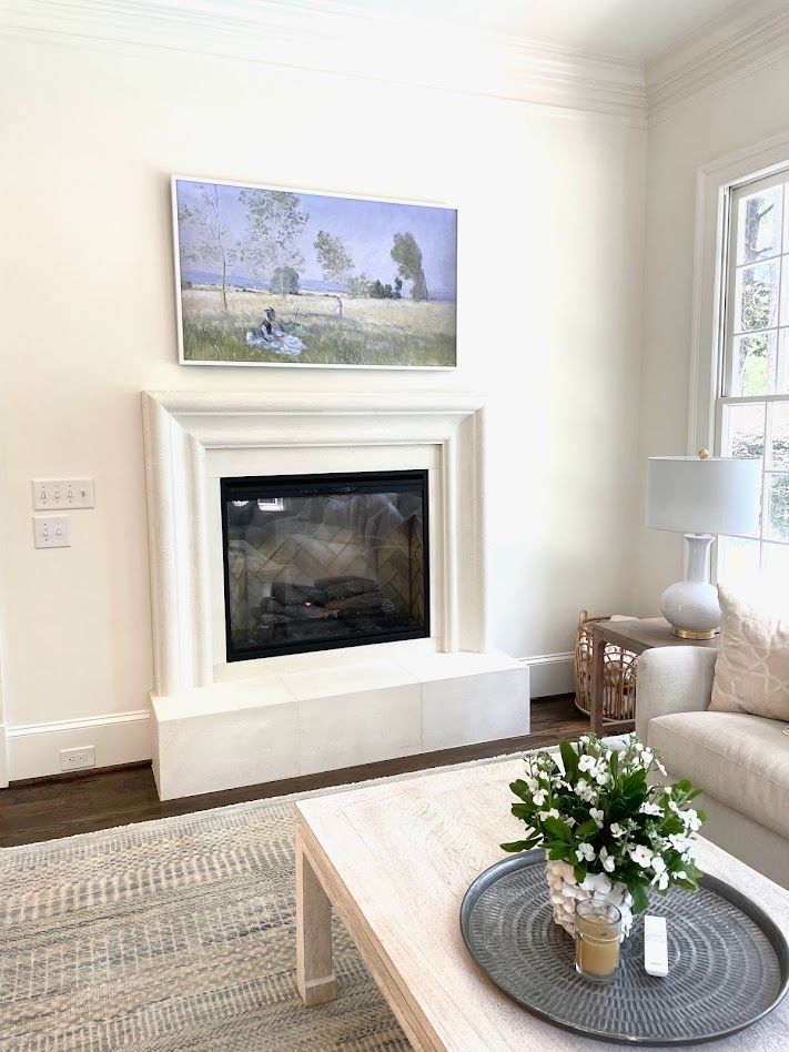 Living room with fireplace, artwork above. Neutral colors: beige, white, grey. Coffee table with flowers, sofa and lamp.
