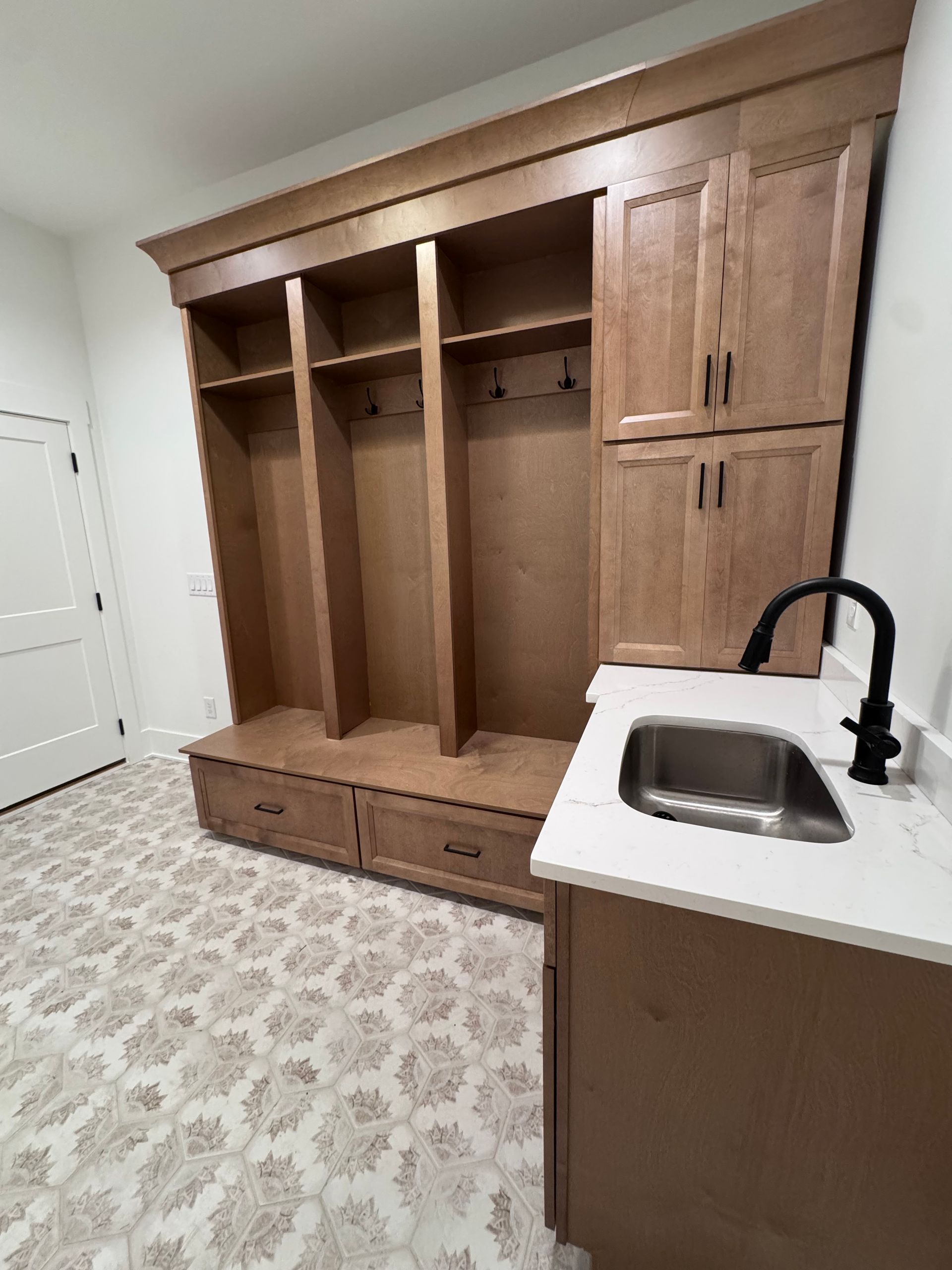 Built-in wooden mudroom storage with lockers, cabinets, and a sink, next to patterned tile flooring.