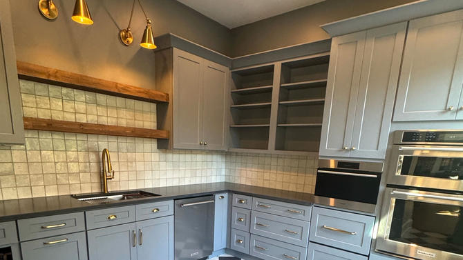 A modern kitchen with grey cabinets, a white tile backsplash, wooden floating shelves, and stainless steel appliances.