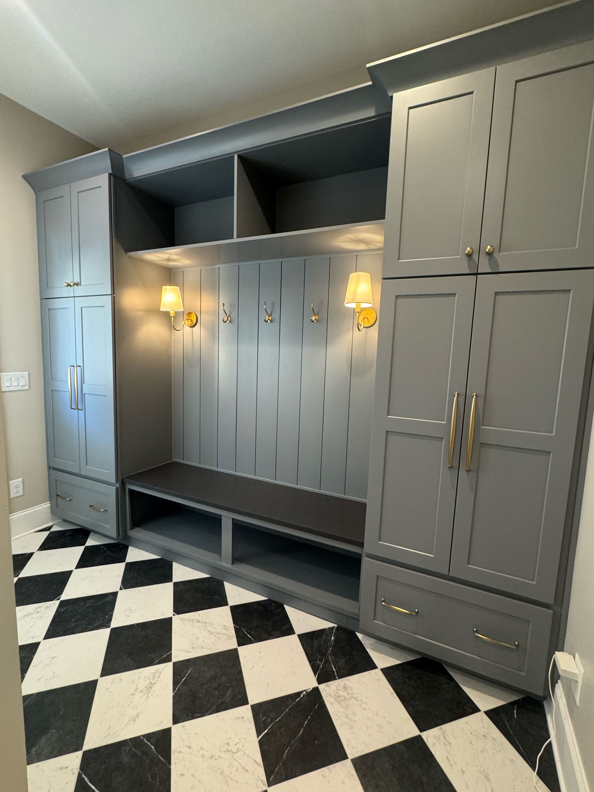 Built-in gray cabinetry with hooks, bench, and shelves. Gold sconces and black/white checkered floor.