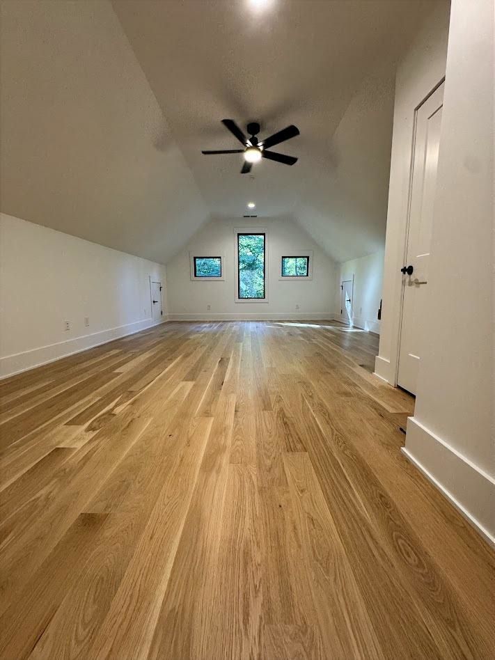 Wooden-floored attic room with white walls, a vaulted ceiling, windows, and a black ceiling fan.