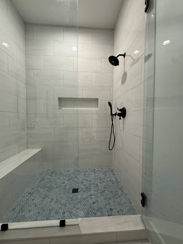 Modern shower with blue mosaic floor, white tiled walls, glass door, bench seat, and black fixtures.