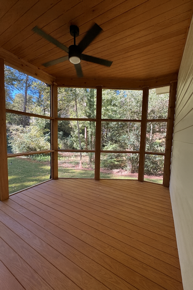 Screened-in porch with brown composite deck and ceiling, black ceiling fan, and view of trees through screens.