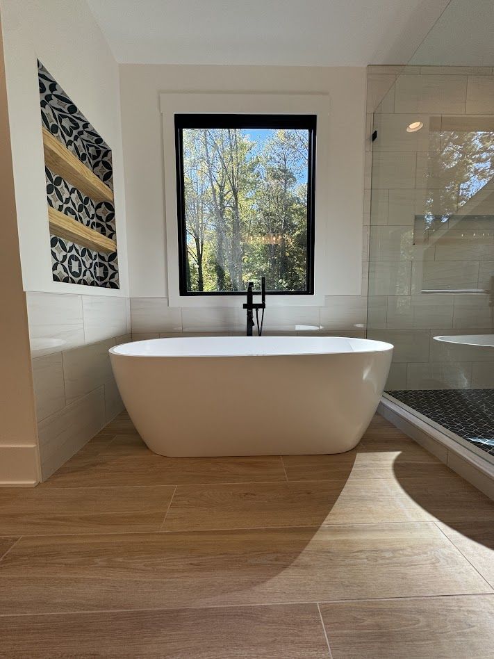 Modern bathroom with a white bathtub, black faucet, window, and wooden flooring.