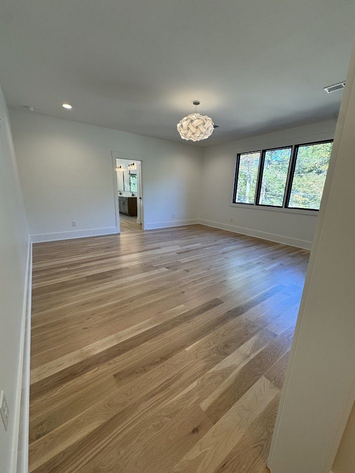 Empty room with hardwood floors, white walls, and a large window. A chandelier hangs from the ceiling.