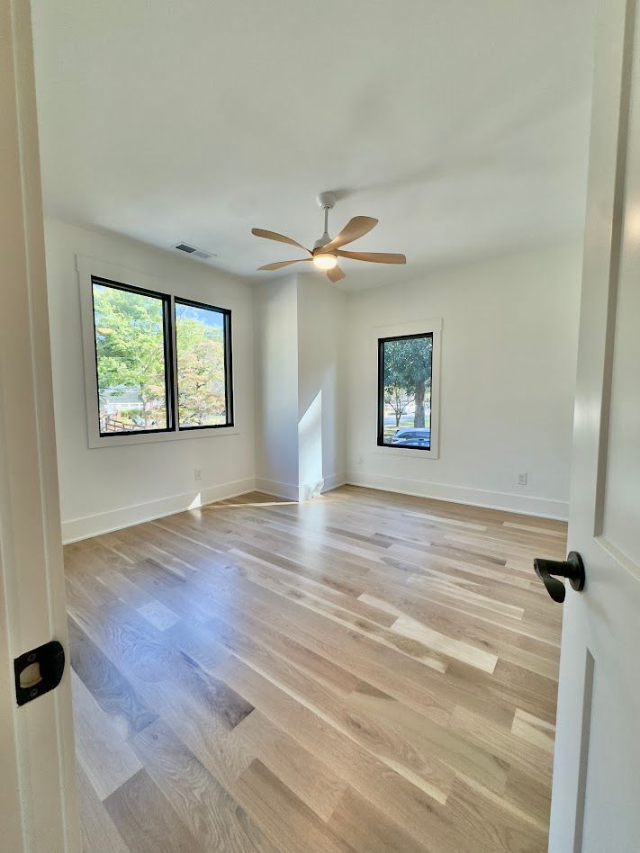 Empty bedroom with hardwood floors, white walls, black-framed windows, and ceiling fan.