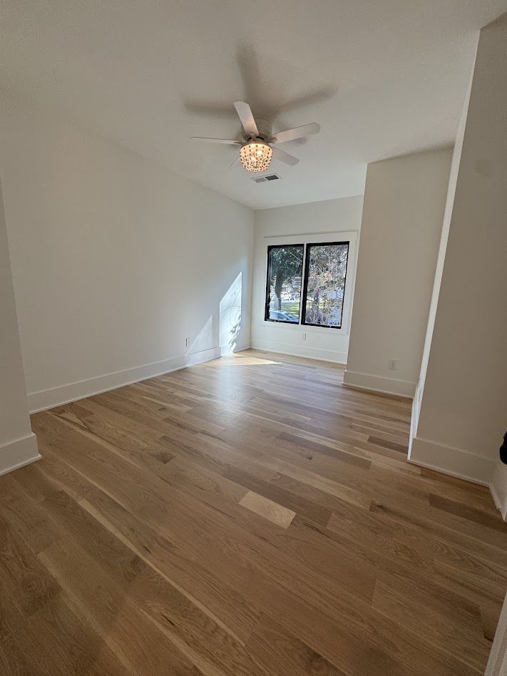 Empty room with hardwood floors, white walls, and a window. A ceiling fan is in the center.
