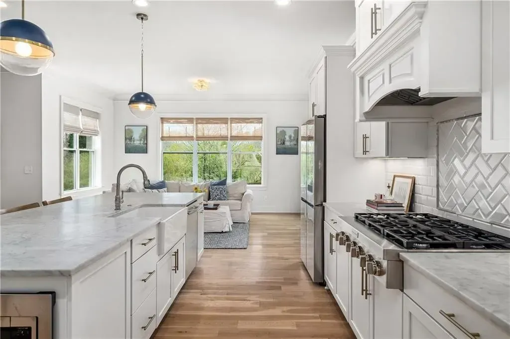 Modern, bright white kitchen with a large marble-topped island, gas stovetop, and view of a living room with wooden floors.