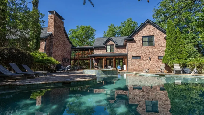 A multi-story brick house with a slate roof features a large backyard swimming pool under a bright blue sky.