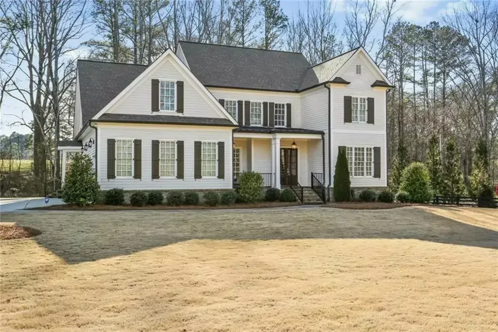 Two-story white house with black shutters, on a brown grass lawn, in front of bare trees.