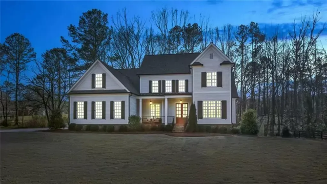 A two-story white house with dark window shutters at dusk, surrounded by tall trees and a lawn.