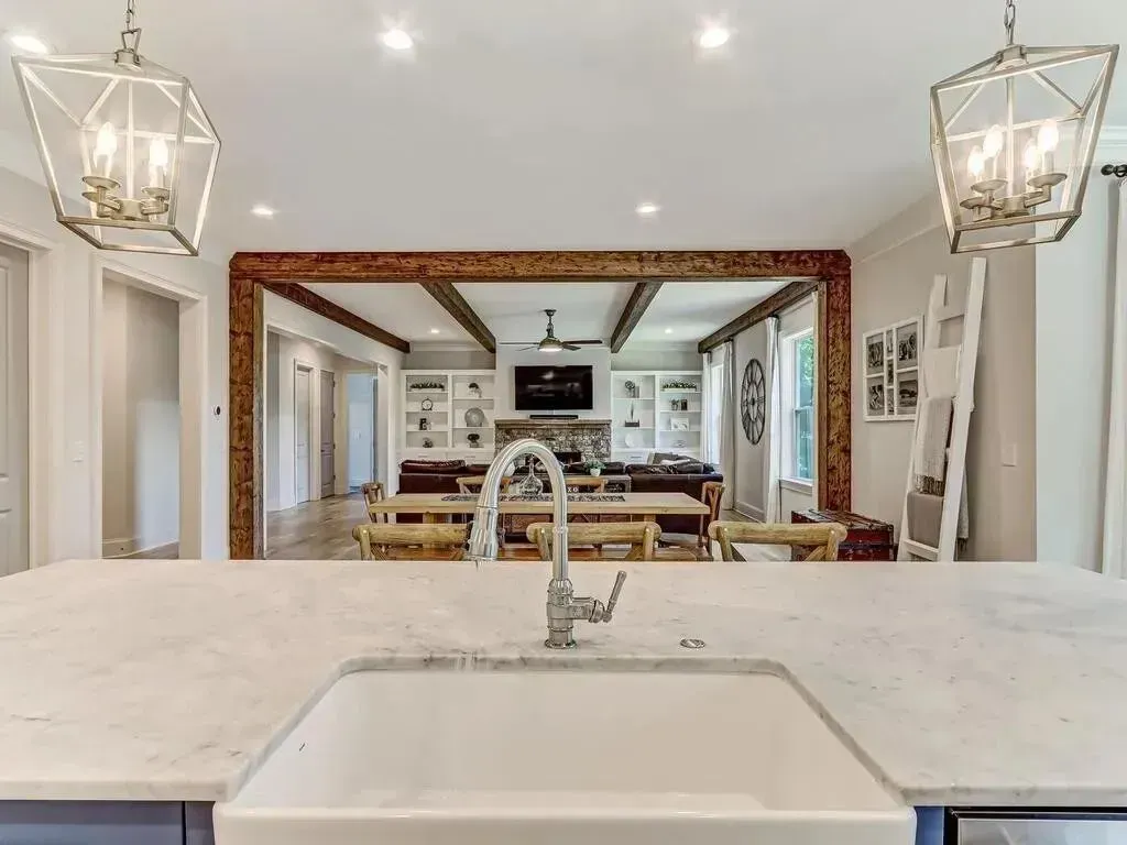 Kitchen sink with marble countertop, looking into living room with wood beams and fireplace.
