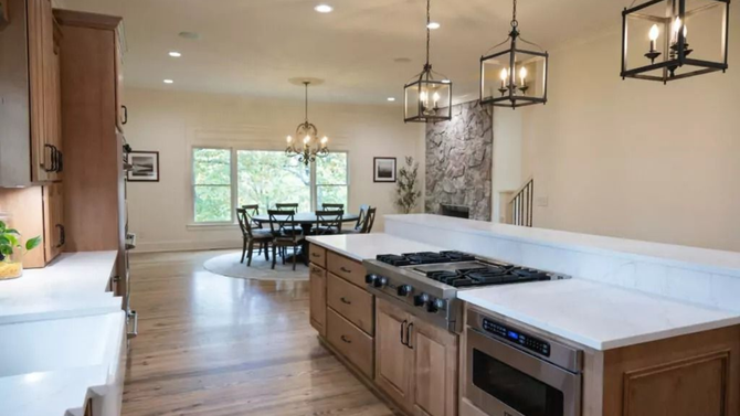 A bright open-concept kitchen with a wooden island, gas stovetop, and dining area featuring a round table and windows.