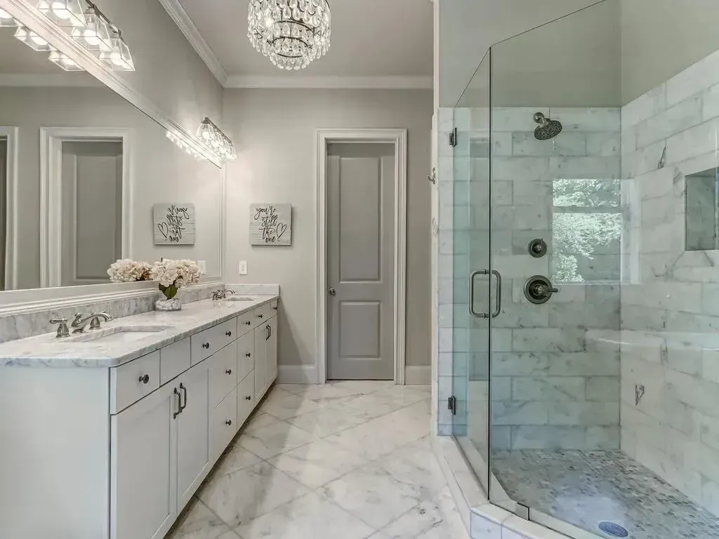 Bathroom with white marble, glass shower, double vanity, and crystal chandeliers.