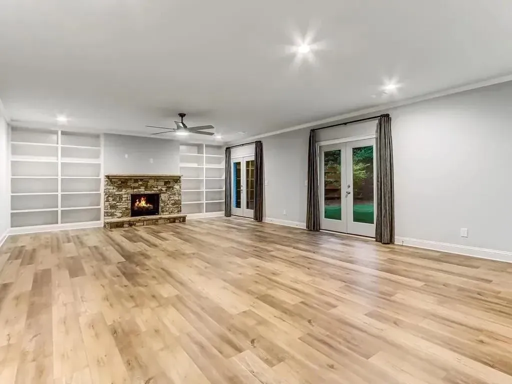 Empty living room with wood flooring, stone fireplace, built-in shelving, and french doors.