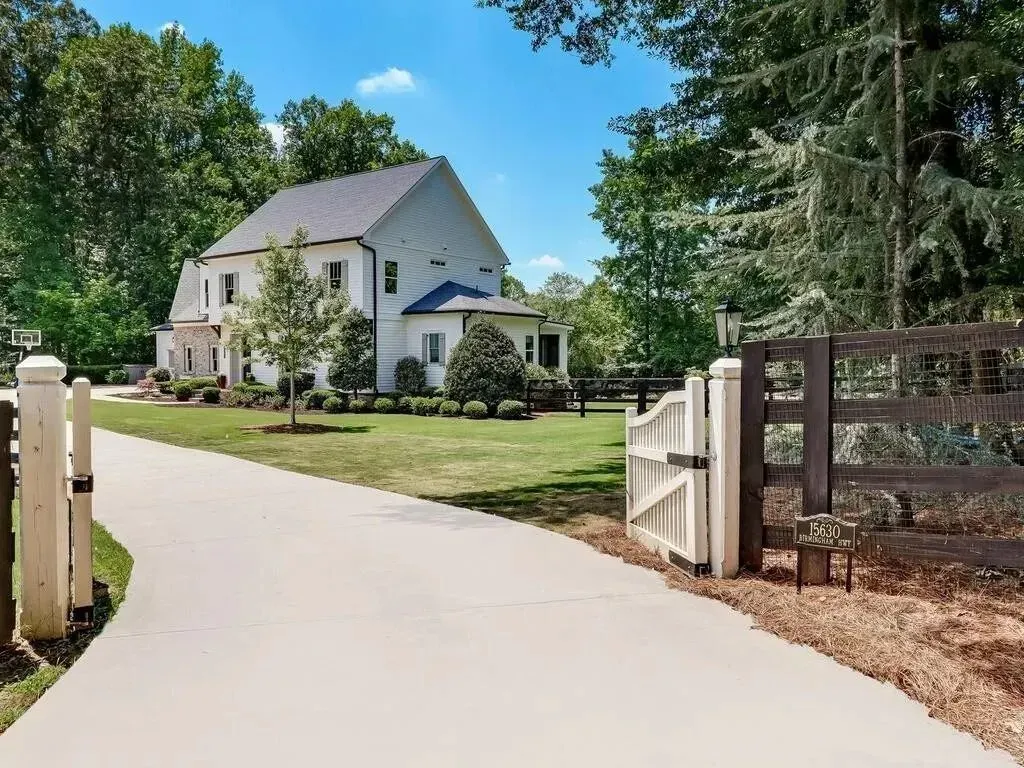 White house with a driveway and gate, surrounded by trees under a blue sky.