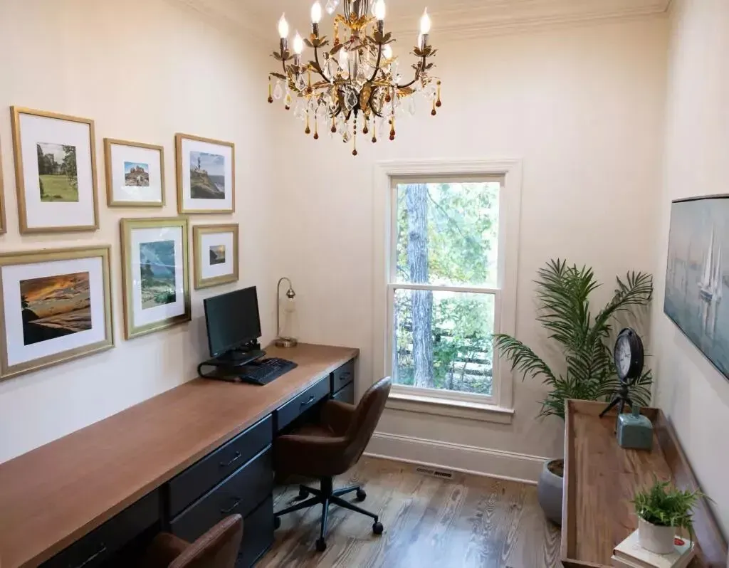 Home office with desk, artwork, chandelier, and a view of trees.