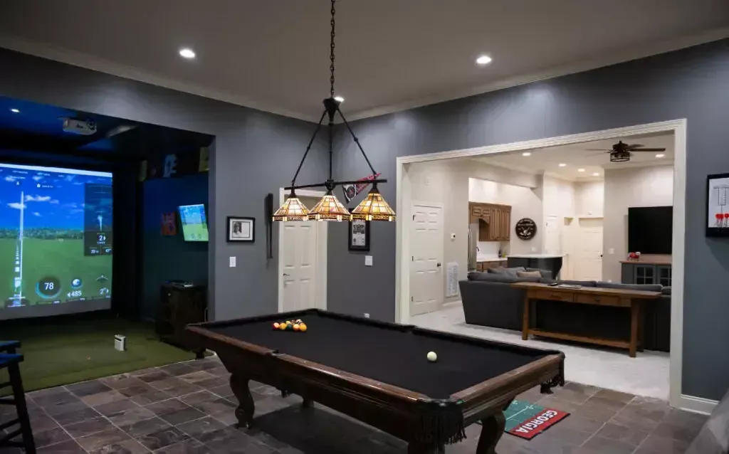 Pool table in a room with a golf simulator. Dark gray walls, decorative lighting, and doorways to other areas.
