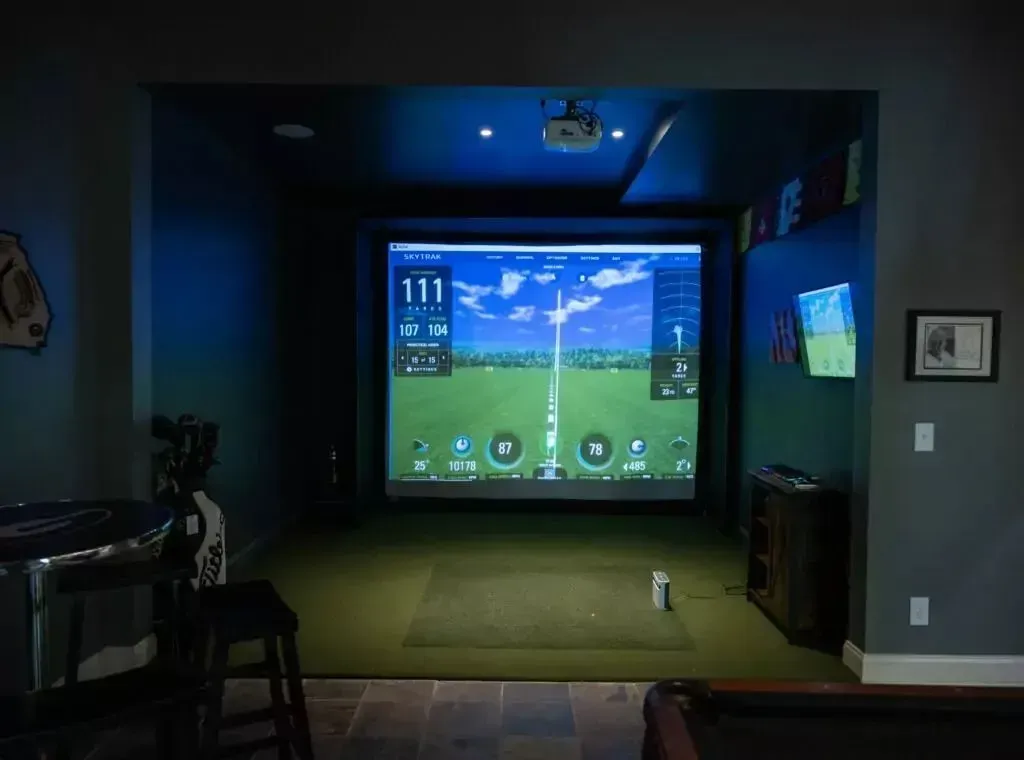 Golf simulator room with screen displaying a golf course, projector, and hitting mat.