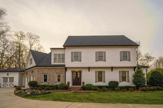 White two-story house with stone and brick exterior, dark roof, and garage on a green lawn.