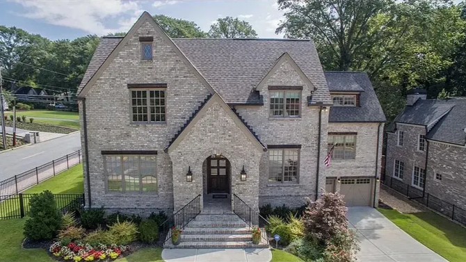 A two-story stone house with a steep roof, central arched entryway, and front yard landscaping under a blue sky.