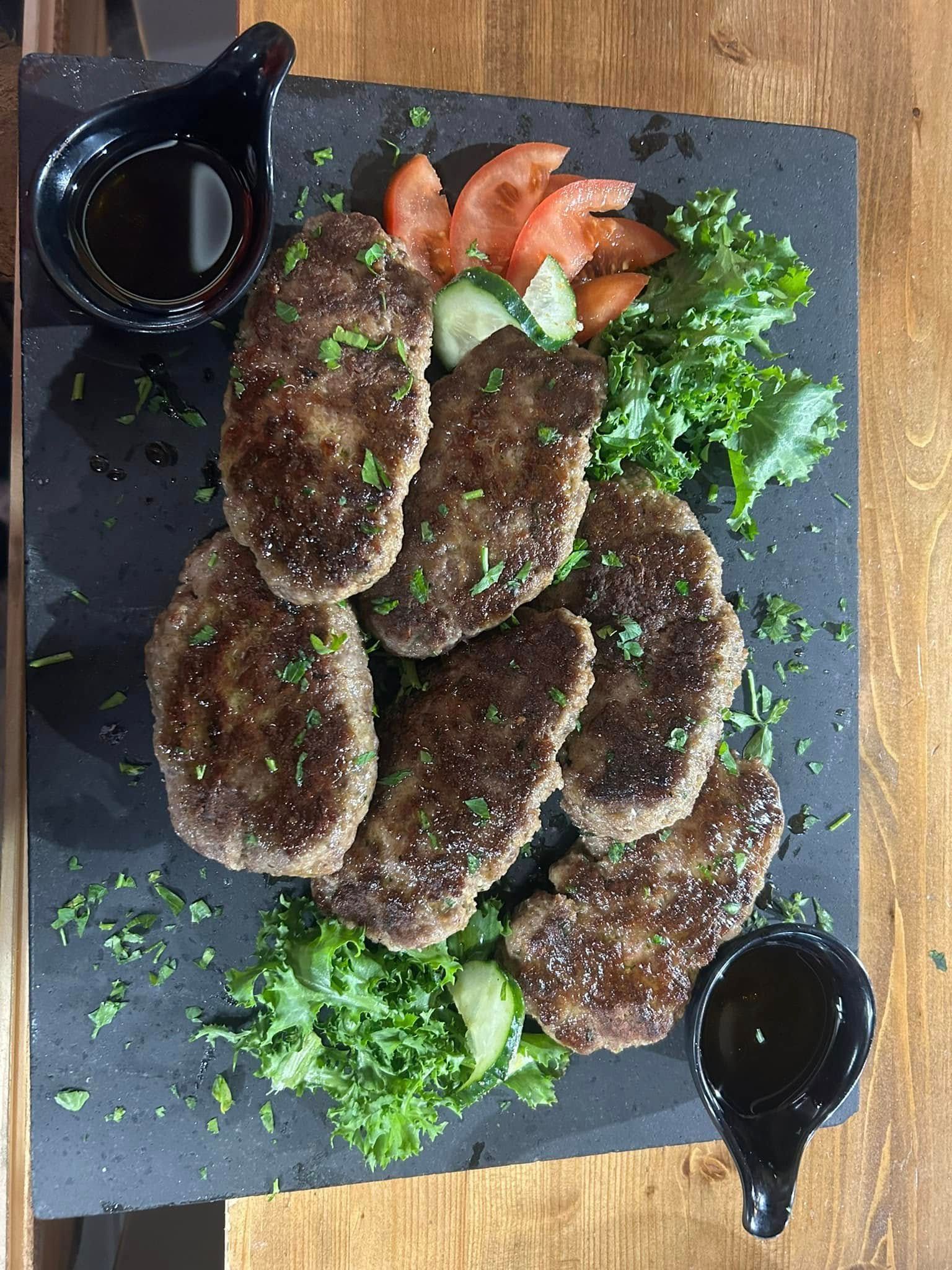 Piatto di polpette di carne alla griglia, guarnite con verdure e pomodori a fette, servite con salse.