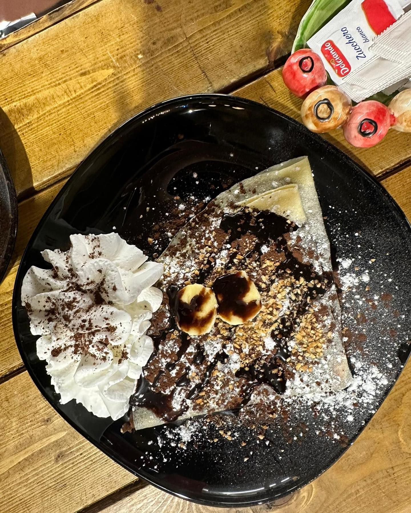 Crêpe con salsa al cioccolato, panna montata, banane e zucchero a velo su un piatto nero, su un tavolo di legno.