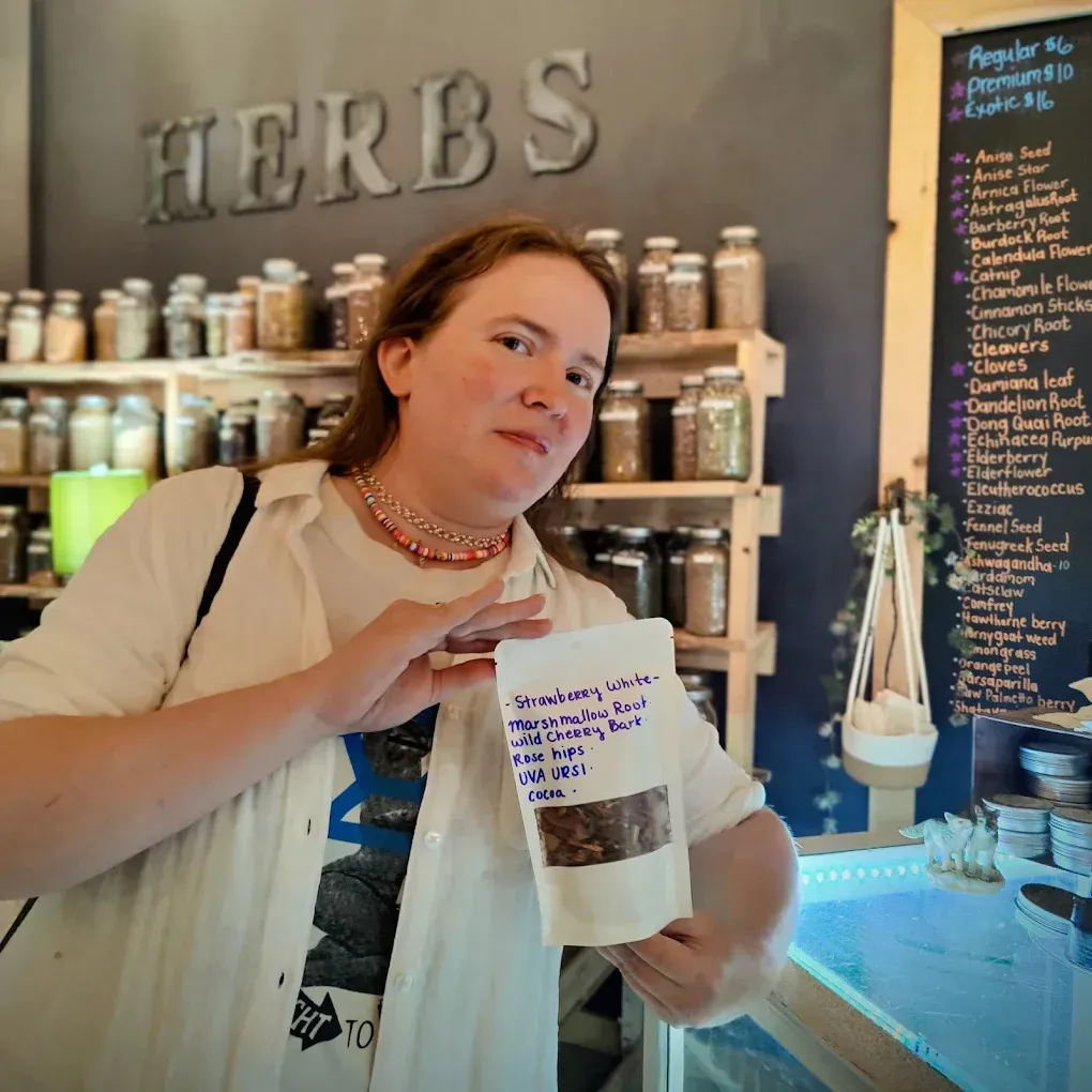 Person holding a bag of herbs inside a shop with shelves of jars and a large HERBS sign. Person holding a bag of herbs inside a shop with shelves of jars and a large HERBS sign.