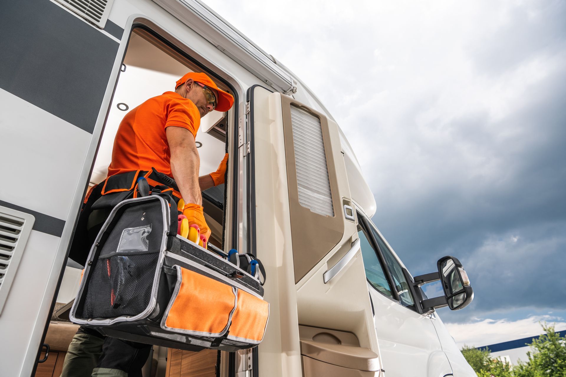 A person in orange work gear exits a white RV carrying a toolbox. Cloudy sky background. A person in orange work gear exits a white RV carrying a toolbox. Cloudy sky background.